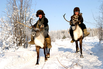 Two Yakut people dressed in warm furry clothes riding reindeer through the snowy forest.