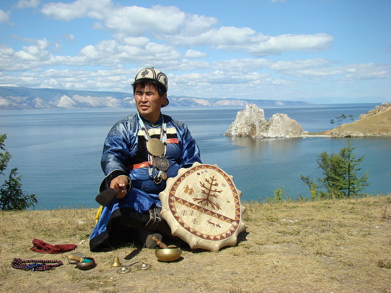 A photo of a Siberian shaman standing on the ledge of a cliff while performing a ritual. Behind him there lies a river and far away beyond it, a chain of hills.