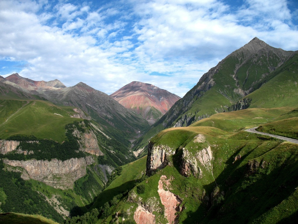 Photo of the Caucasus Mountains, a valley is splitting the rocky formations covered by meadow, under a cloudy sky.